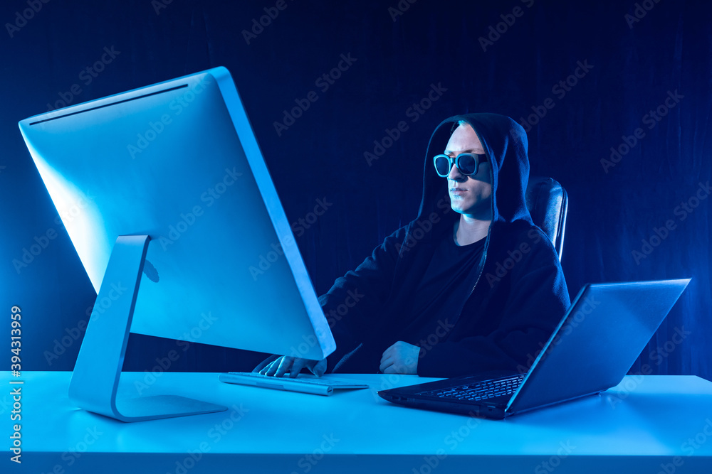 Poster hacker man. hacker is looking at monitor. hacker in sunglasses and a hood. young man near a computer