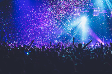 Fototapeta na wymiar A crowded concert hall with scene stage lights, rock show performance, with people silhouette, colourful confetti explosion fired on dance floor air during a concert festival