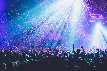 Fototapeta na wymiar A crowded concert hall with scene stage lights, rock show performance, with people silhouette, colourful confetti explosion fired on dance floor air during a concert festival