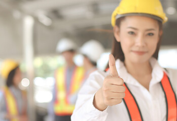 confident Asian Female architect or engineer  holding blue print paper, standing in front of her team smiling , looking at camera and showing her thumb up.Selective focus on woman's thumb.
