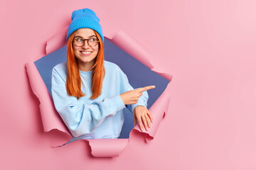 Smiling ginger woman feels optimistic points away on blank space shows advertisement wears blue hat...