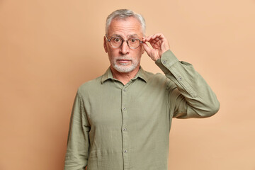 Surprised grey haired handsome man keeps hand on rim of spectacles hears amazing news wears formal...
