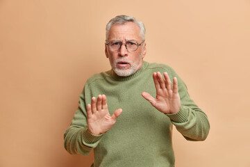 Studio shot of scared grey haired man keeps palms in defensive gesture asks not to come closer sees...