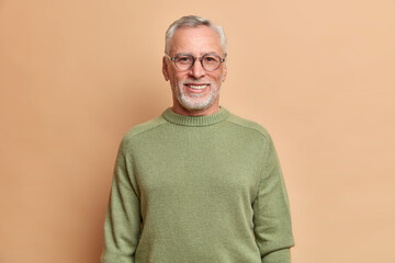 Half length shot of cheerful senior man smiles happily with white teeth wears optical glasses and sweater isolated over brown background. Happy grandfather glad grandchildren came to visit him
