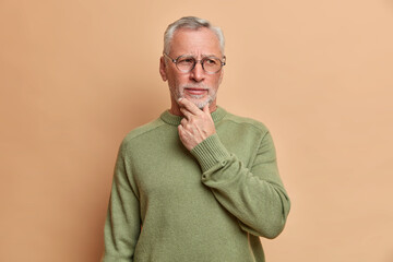 Pensive serious old bearded man holds chin and looks away thoughtfully thinks over received...