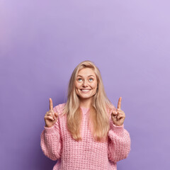 Happy glad European woman with blonde hair smiles pleasantly indicates above on blank space gives place for your advertisement wears knitted sweater isolated over purple background. Check this out