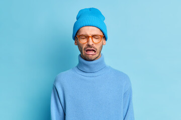 Depressed crying man being in stressful situation stands desperate as faces problem feels regret blames himself in failure wears trendy blue hat turtleneck spectacles poses indoor. Pessimistic person