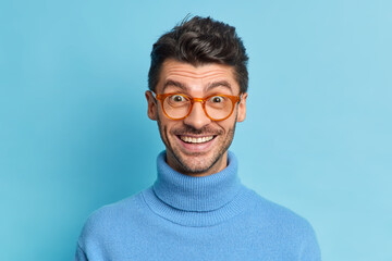 Headshot of glad hipster guy with bristle smiles broadly reacts on positive news hears something excellent wears optical glasses casual turtleneck isolated over blue background. Good emotions