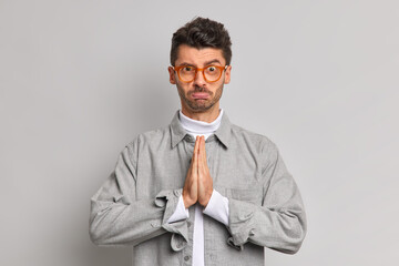 Upset desperate unshaven European man keeps palms pressed together clasps hands purses lower lips wears glasses and grey shirt asks to give him one more chance apologizes for doing something wrong