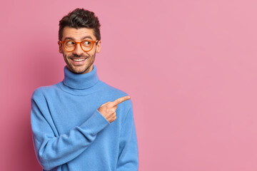 Use this copy space wisely. Glad handsome man in casual outfit points away on right advetises something smiles happily wears blue turtleneck isolated over pink background shows good offer to you