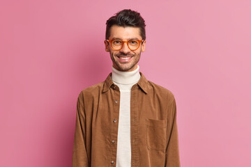 Obraz premium Handsome cheerful European guy with thick bristle smiles broadly wears optical spectacles and brown shirt being in good mood dresses for work poses against pink background. Happy male student