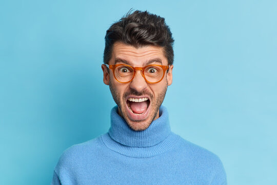 Close up shot of outraged irritated man keeps mouth widely opened screams with annoynace expresses negative emotions wears spectacles and poloneck poses against blue background. Negative emotions