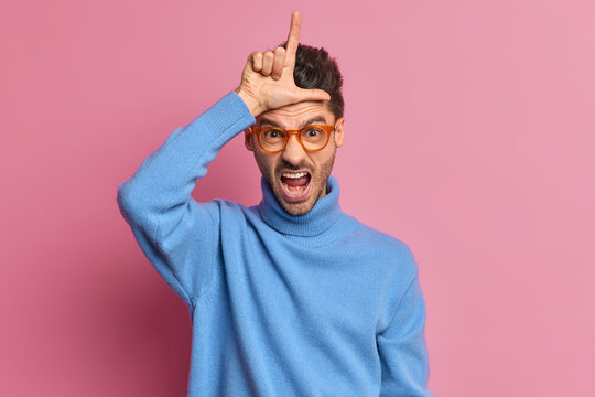 Annoyed adult man makes looser gesture wears spectacles and blue turtleneck has failure or unlucky day as lost his job upset about dismissal makes L finger sign over forehead isolated on pink wall