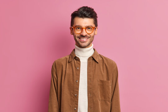 Handsome cheerful European guy with thick bristle smiles broadly wears optical spectacles and brown shirt being in good mood dresses for work poses against pink background. Happy male student