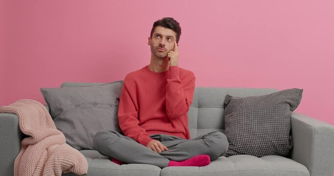Serious young European man sits thoughtful keeps index finger on temple thinks deeply and got excellent idea poses on sofa isolated over pink background recalls necessary information in mind