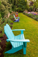A long perspective view of an English style garden with a row of colorful Adirondack benches flanked by beds of mixed flowers - Shallow field of focus