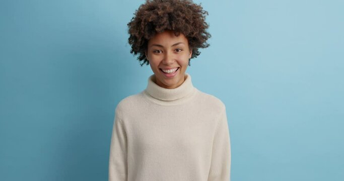 Serious dark skinned woman asks what then nods head and says yes smiles broadly feels very happy has pleasant talk with someone wears casual sweater isolated on blue background. Human face expressions
