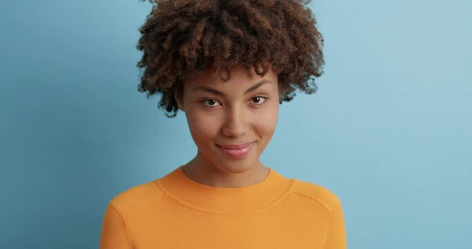 Young Afro American woman looks at camera with satisfied expression winks eye and flirts has mysterious gaze and happy smile poses against blue background sends positive vibes cheers friend up