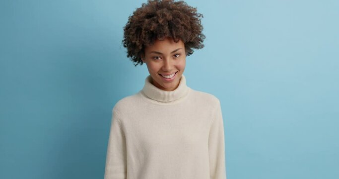 Smiling glad dark skinned woman with curly hair smiles pleasantly and winks eye flirts to handsome boyfriend wears casual white sweater looks happily at camera models against blue background