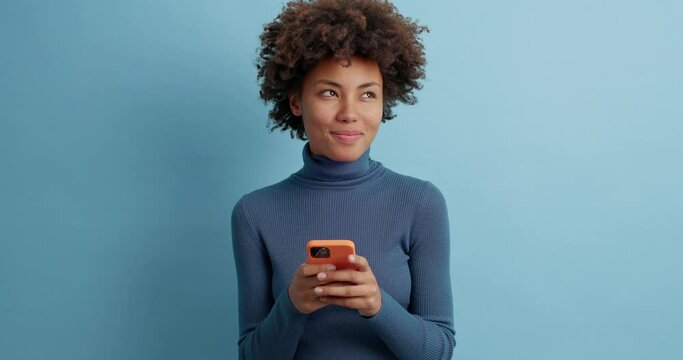 Cheerful curly haired young female in casual turtleneck types message in hurry uses smartphone for online communication enjoys browsing social media poses against blue background reads received sms