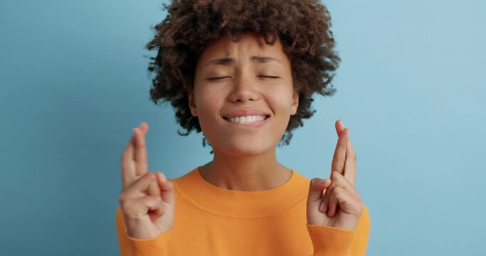 Hopeful Afro American woman crosses fingers for good luck truly believes her dreams come true waits for important results stands intensed dressed in orange jumper isolated over blue background