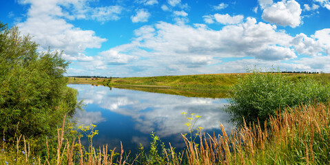 Beautiful panoramic rural landscape with calm river and green hills with trees at sunny summer day.