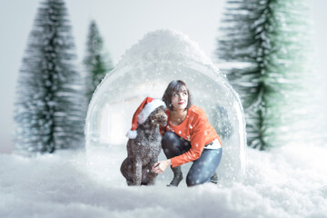 Caucasian woman wearing a christmas sweater and her dog with santa claus hat inside a snow globe. Concept of coronavirus quarantine during christmas season