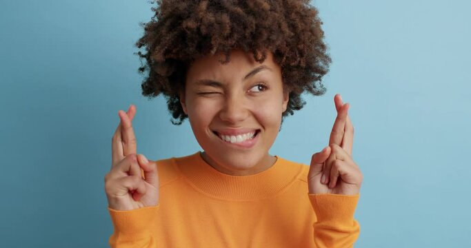 Beautiful hopeful dark skinned woman crosses fingers for good luck smiles broadly and anticipates for something good happen or miracle poses against blue background. Hopes and dreams concept