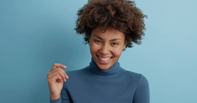 Happy sincere Afro American woman laughs out from joy hears hilarious joke smiles broadly dressed in casual turtleneck isolated over blue background. Positive emotions and feelings. Close footage