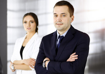 Portrait of a friendly middle aged businessman in a dark blue suit, standing with crossed arms together with a colleague in a sunny modern office. Modern business concept