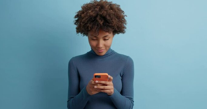 Happy Afro American woman exchanges lovely messages with boyfriend holds mobile phone types and browses in network smiles pleasantly dressed in casual turtleneck isolated over blue studio wall