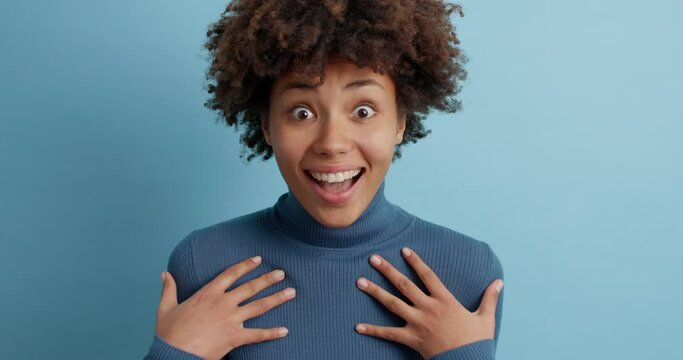 Beautiful young Afro American woman looks with surprised face expression at camera and says wow impressed touched to hear something excellent poses against blue background. Reaction concept.
