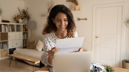 Happy young Caucasian girl sit at desk at home reading good news in postal letter correspondence. Smiling millennial female receive get pleasant message or promotion notice in post paperwork.