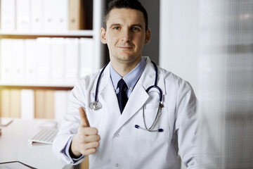 Friendly male doctor sitting and showing OK sign with thumbs up at his working place in sunny clinic. Perfect medical service in hospital