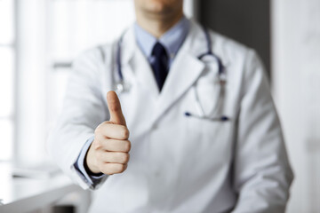 Unknown male doctor sitting with thumbs up sign at his working place in clinic, closeup. Perfect medical service in hospital. Medicine and healthcare concept