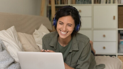 Smiling young Caucasian woman in headphones laugh look at laptop screen have pleasant webcam digital virtual conference. Happy female in earphones talk speak on video call on computer at home.