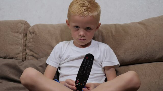 Close-up of a sad little blond boy sitting on the sofa at home, holding a remote control. The child is punished and does not watch TV.