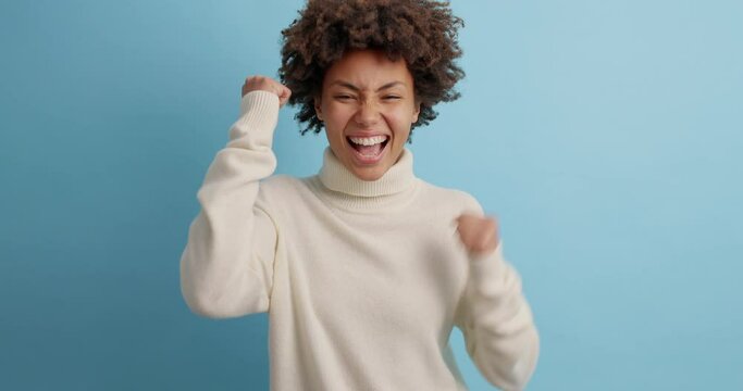 Overjoyed curly haired woman wins competition achieves goal and makes triumph gesture says yes feels like winner shows fist bump happy dream come true wears white jumper poses against blue wall