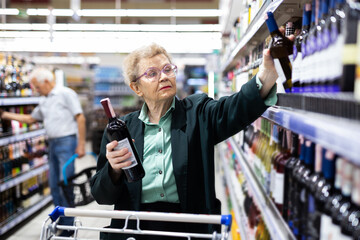 mature woman with glasses chooses bottle of wine in alcohol section of supermarket