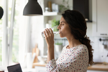 Young Caucasian woman stand at home kitchen drink clean pure still mineral water from glass for...