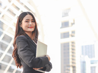 Fototapeta premium Young Asian business woman wearing black suit, holding computer laptop, smiling and looking at camera. Outdoor with business building background.