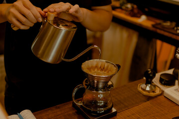 Black shirt barista pouring hot water on coffee ground with filter from silver teapot to transparent chrome drip maker on wooden table cafe shop. Drip brewing, filtered coffee.
