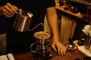 Fototapeta na wymiar Black shirt barista pouring hot water on coffee ground with filter from silver teapot to transparent chrome drip maker on wooden table cafe shop. Drip brewing, filtered coffee. 