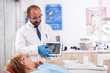 Stomatolog in uniform during consultation of senior woman explaining diagnostic. Medical teeth care taker holding patient radiography on tablet pc near patient standing up.