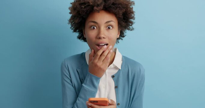 Young shocked curly haired young woman feels surpsied looks with disbelief at camera points at smartphone display fascinated by exciting news or received commets on her blog post. Omg look there