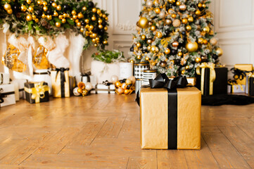 gold gift box with black ribbon on wooden floor in room with Christmas decor in gold and black colors : Christmas tree with balls and garlands and many Packed gifts under it, selective focus