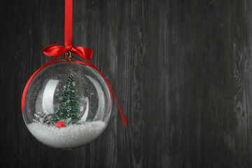 Beautiful Christmas snow globe hanging on black wooden background, space for text