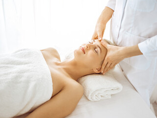 Beautiful blonde woman enjoying facial massage with closed eyes in sunny spa center. Relaxing treatment in medicine and beauty concepts
