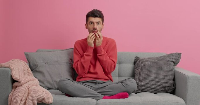 Scared young man watches horror film with special effects reacts on scary scene stares frightened at camera feels appalled poses alone on comfortable sofa. Thrilling film moment. Guy views tv thriller