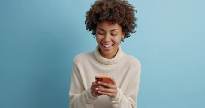 Young happy curly haired woman checks message on smartphone reads comments under post on her webpage dressed in casual white sweater poses against blue background checks information in internet